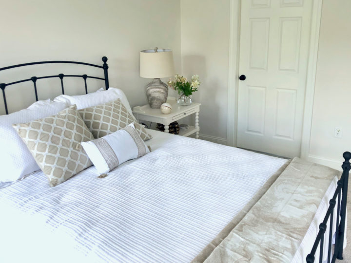 Neutral transitional guest bedroom with black metal panel bed, white mattelasse coverlet, textured geo print window panels, resin table lamps, and white open nightstand.