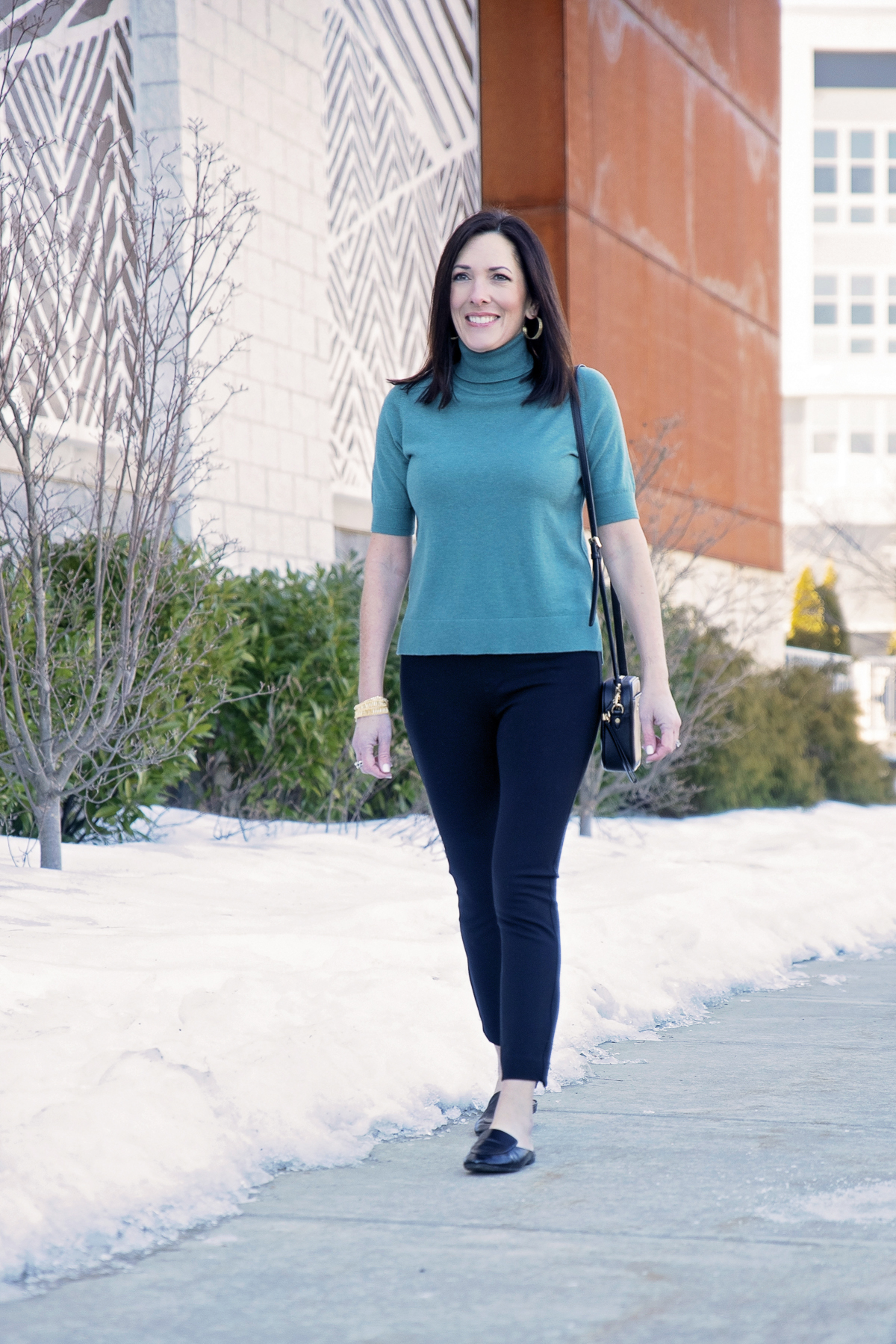 Jo-Lynne Shane wearing a polished spring work wear look with a teal short-sleeve turtleneck, black Spanx skinny pants, and Everlane loafer mules.